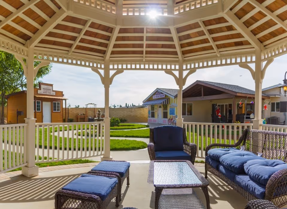 Gazebo seating area with wicker furniture and blue cushions overlooking a grassy courtyard and small outbuildings.