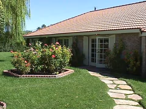 A single-story building with a tiled roof, a front door, and flowering shrubs along a walkway.