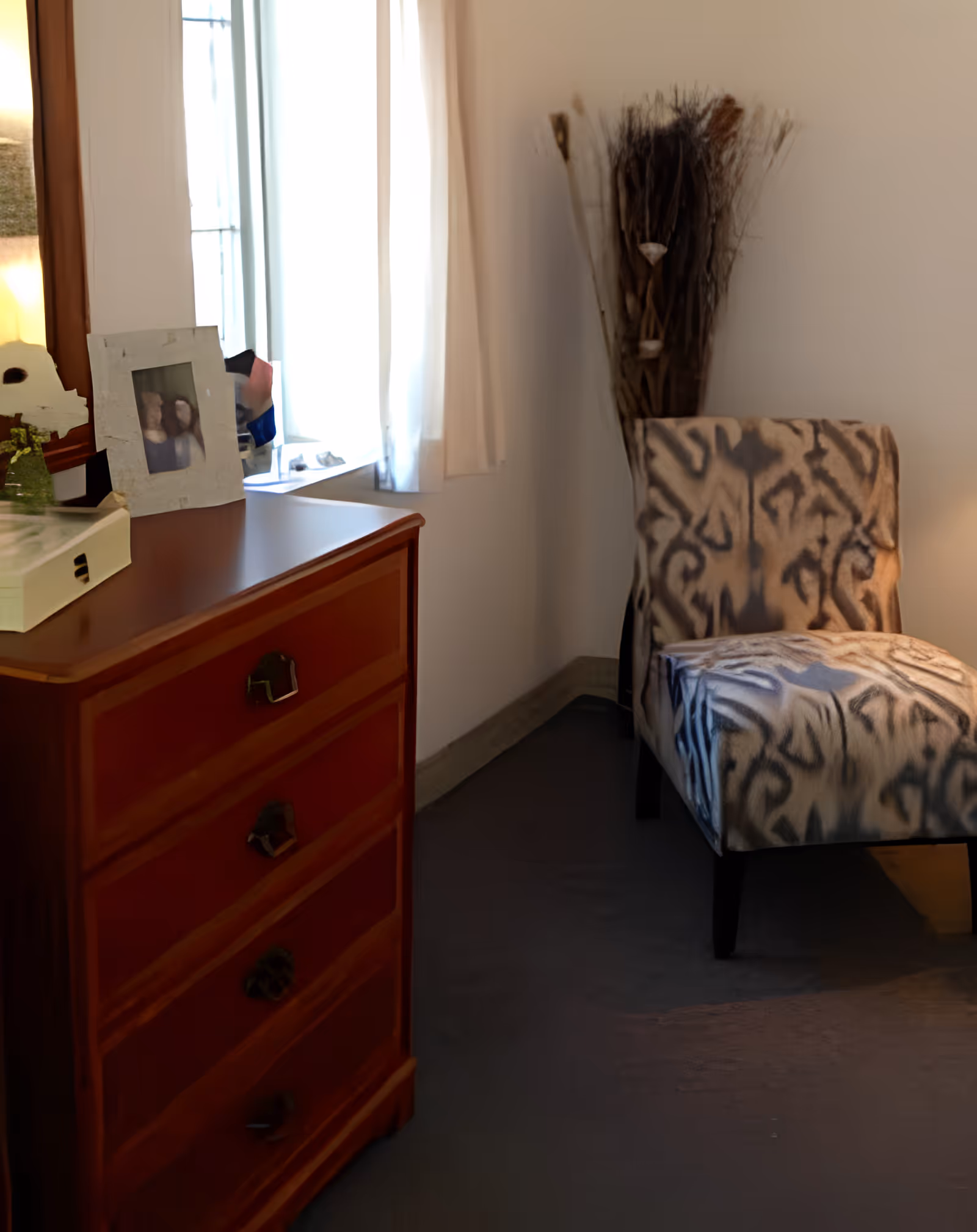 A small sitting area with a wooden chest of drawers topped with a framed photo and a patterned upholstered chair by a window.