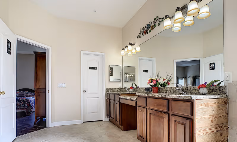 A bathroom area with a large mirror above a granite countertop featuring two sinks and wooden cabinets underneath. There are two doors visible, one leading to a bedroom with a bed and wooden furniture, and the other door closed. The bathroom has beige walls and tiled floors, with decorative lighting fixtures above the mirror and a floral arrangement on the countertop.