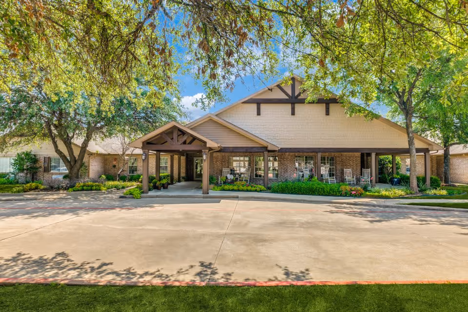 Front exterior view of a single-story senior living facility building with a covered entrance, surrounded by trees and greenery under a clear blue sky.