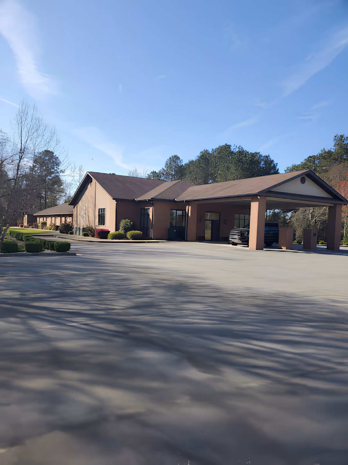 Exterior view of a single-story senior care facility building with a covered entrance and a driveway in front, surrounded by trees and bushes under a clear blue sky.