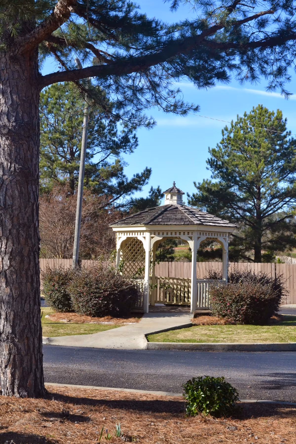 A white wooden gazebo with a shingled roof is situated in a landscaped outdoor area surrounded by bushes and pine trees under a clear blue sky.
