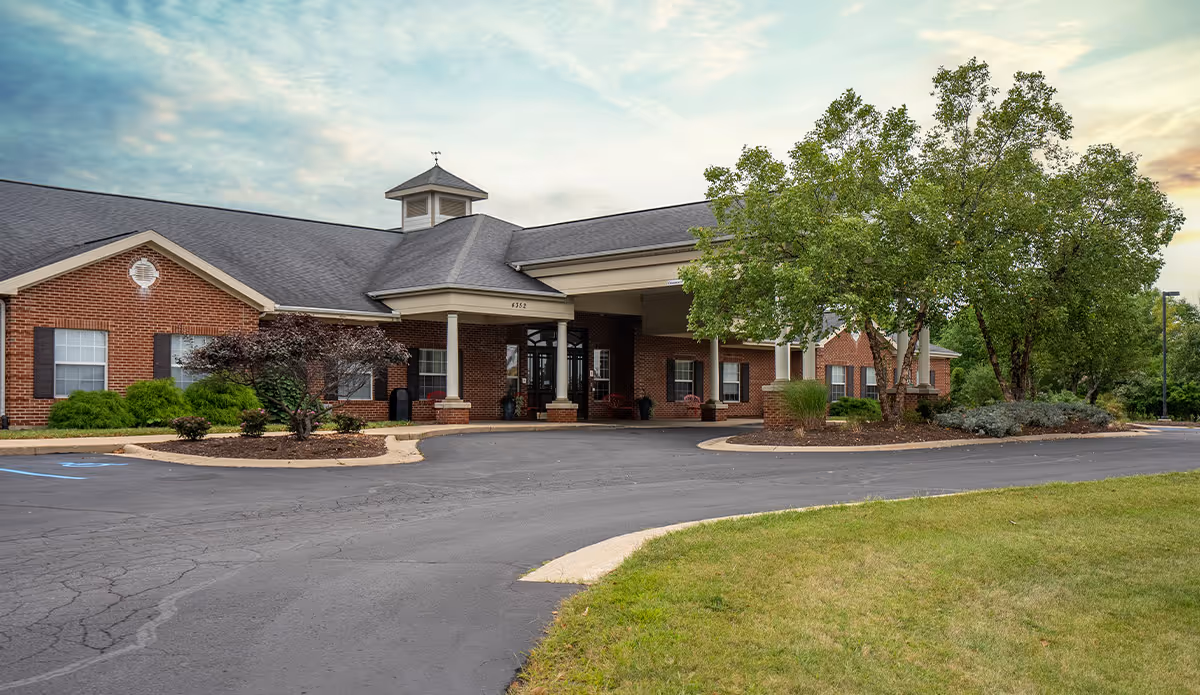 Exterior view of a single-story brick building with a covered entrance, surrounded by trees and landscaping under a partly cloudy sky.