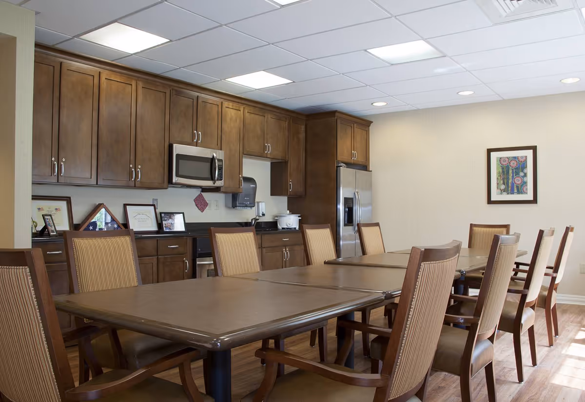 Long dining table with multiple chairs in a communal dining area and kitchenette with wooden cabinets and stainless appliances.