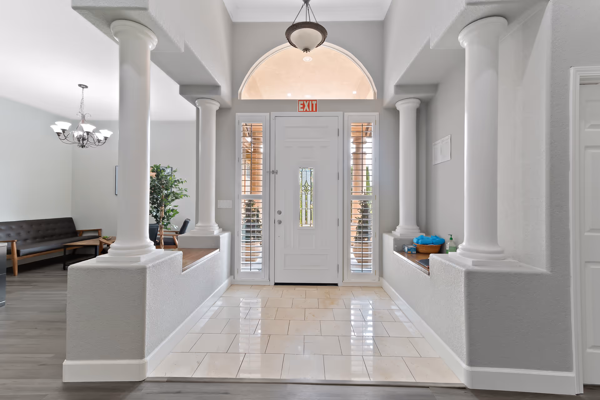 Bright entrance hallway with white tiled floor, white front door with glass panels, and decorative columns on both sides. To the left, a seating area with a dark cushioned bench, wooden chairs, a coffee table, and a chandelier overhead. A potted plant is also visible near the seating area.