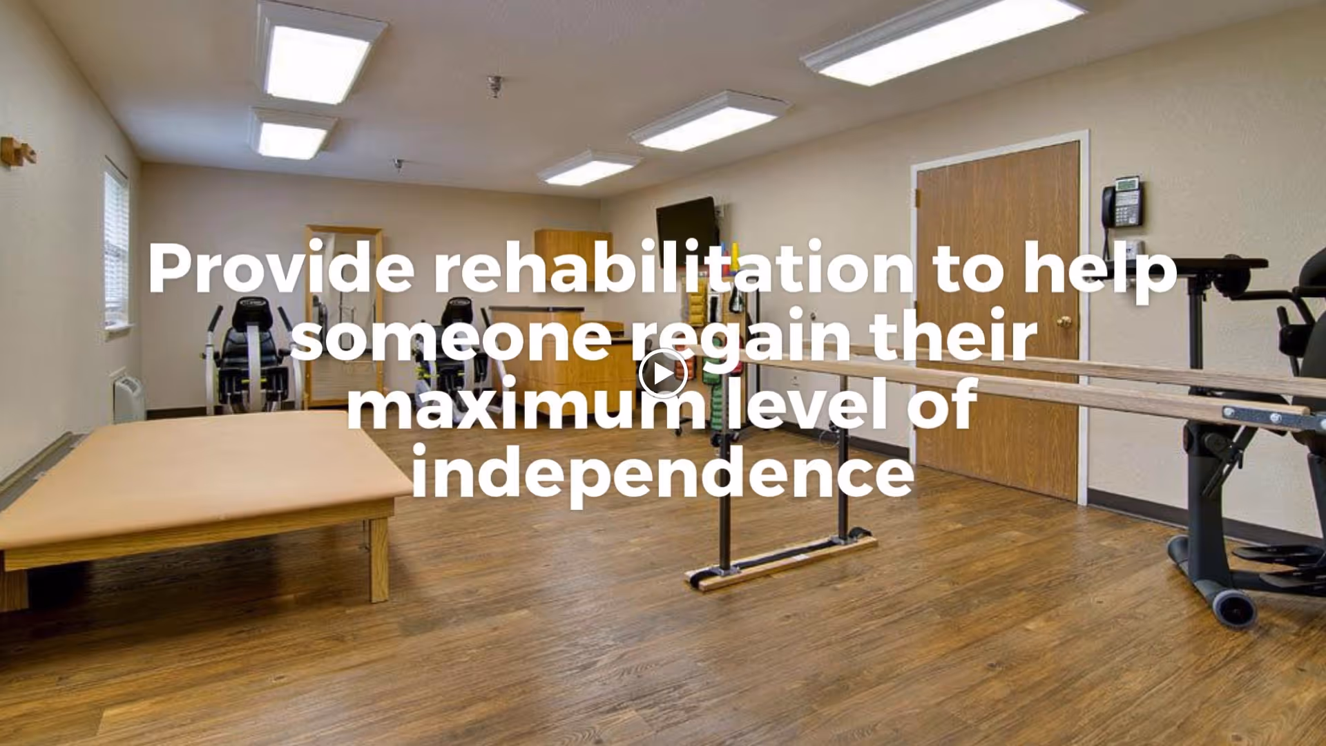 A rehabilitation room with wooden flooring, parallel bars for walking exercises, exercise bikes, and a padded therapy table. The room has beige walls, fluorescent ceiling lights, a wooden door, and a wall-mounted TV.