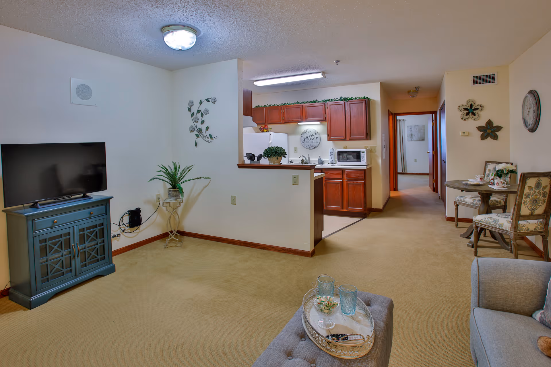 Interior view of a senior living facility apartment showing a living room area with a TV on a blue cabinet, a small plant on a stand, and a cushioned ottoman with a tray holding glasses and a bowl. The kitchen is visible in the background with wooden cabinets, a microwave, and a refrigerator. To the right, there is a small round dining table with two chairs and decorative wall hangings. A hallway leads to other rooms.