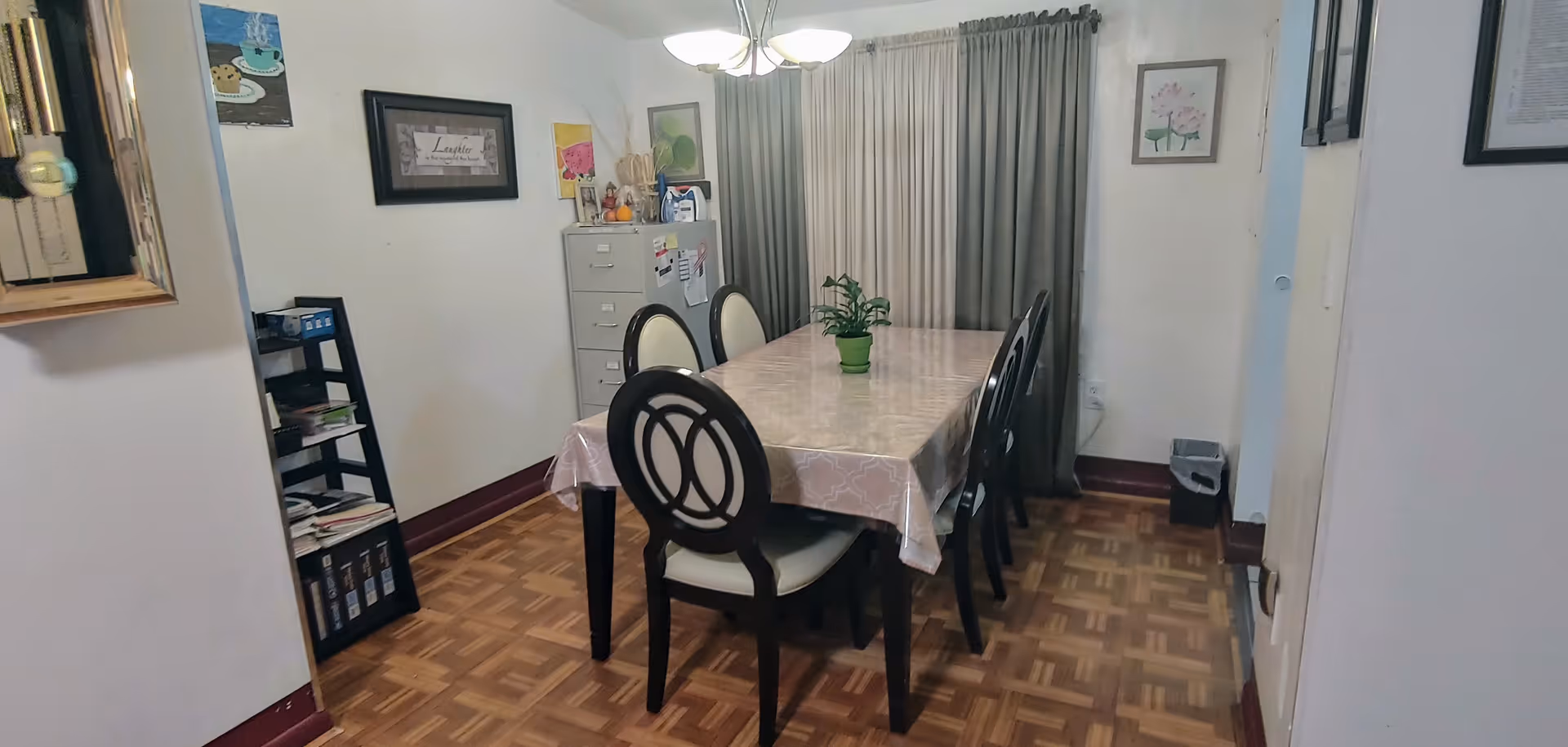 Dining room with a rectangular table covered by a patterned tablecloth, six chairs, a small potted plant, a filing cabinet and closed curtains.