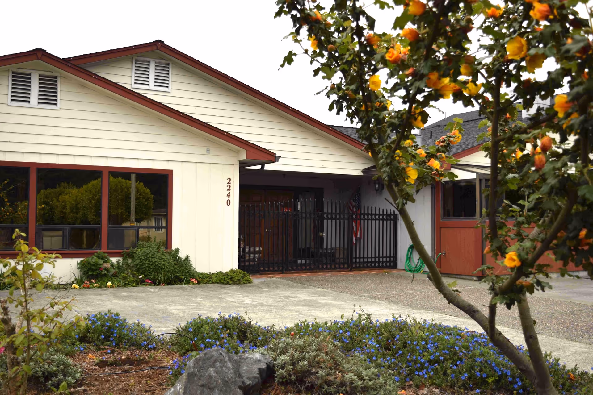 Exterior view of Frye's Care Home showing a single-story building with cream-colored siding and red trim. The building has large windows and a gated entrance. In the foreground, there are flowering plants and a tree with yellow and orange blossoms.