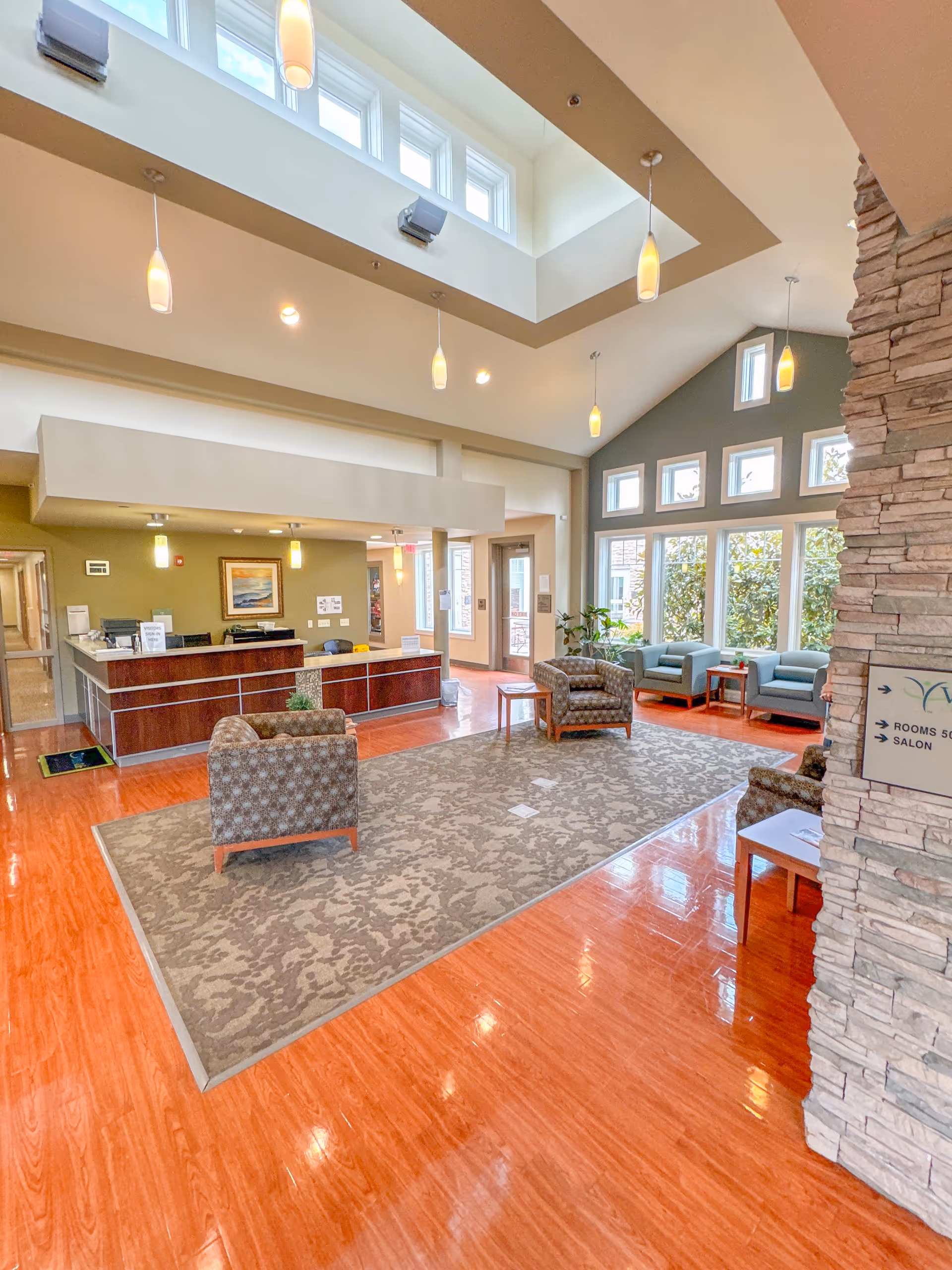 Bright and spacious senior living facility lobby with high ceilings and multiple pendant lights. The area features a reception desk, several armchairs arranged on a large patterned rug, and large windows letting in natural light. A stone pillar with a directional sign is visible on the right side.