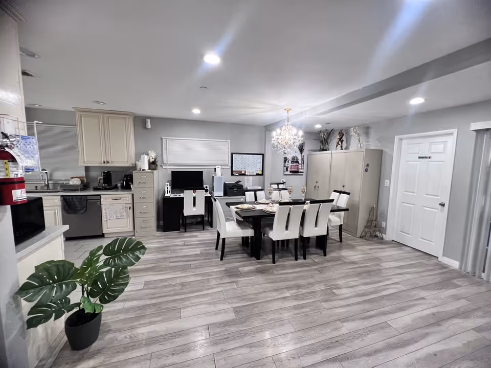Open-plan interior showing a dining table with white chairs beneath a chandelier next to a kitchenette and cabinets.