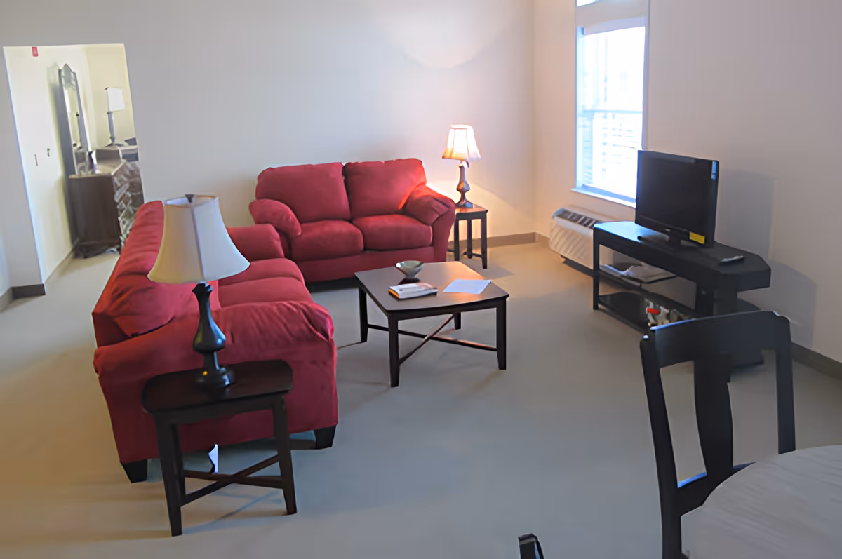 A living room with two red sofas, a wooden coffee table with some papers and a bowl on it, two side tables each with a lamp, a TV on a black stand near a window, and a dining table with chairs partially visible in the foreground.