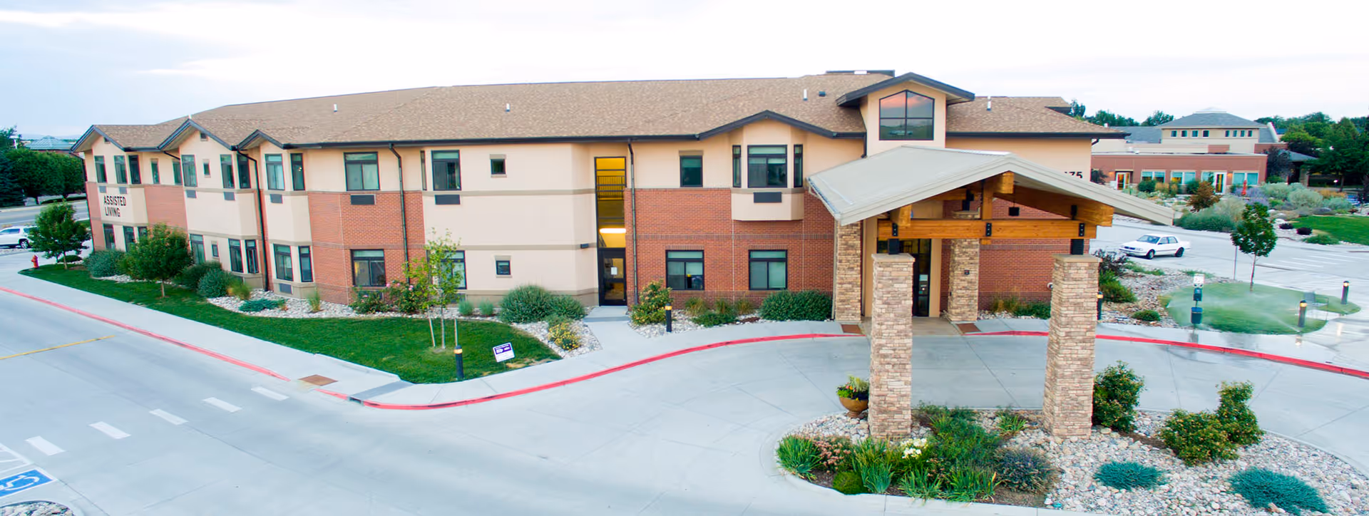 Exterior view of Columbine Commons Assisted Living facility showing a two-story building with a covered entrance supported by stone pillars, surrounded by landscaped greenery and a parking area.