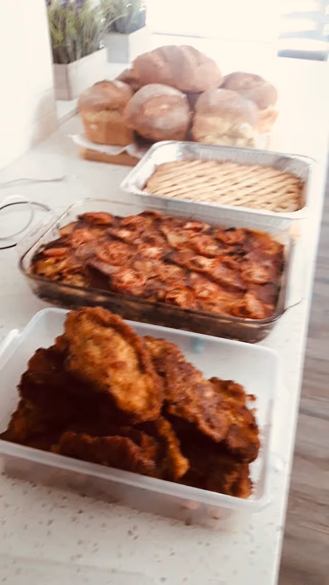 A kitchen countertop with various homemade foods including a container of fried chicken, a glass baking dish with a tomato-topped casserole, a foil tray with a lattice-topped pie, and a stack of bread loaves in the background.