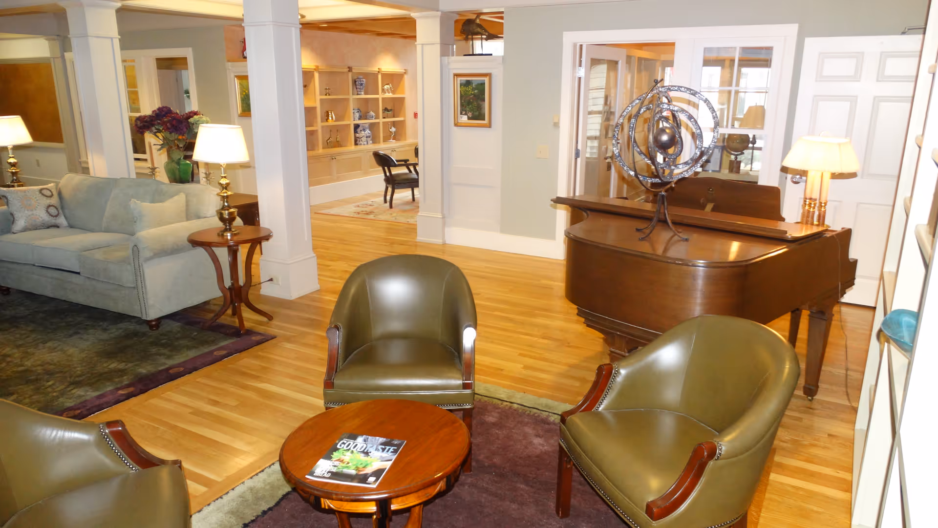 A cozy living room area with three green leather armchairs arranged around a small round wooden table with a magazine on it. There is a light blue sofa with pillows, two table lamps on wooden side tables, a grand piano with a decorative metal sculpture on top, and built-in shelves with decorative items in the background. The room has wooden flooring and soft lighting.