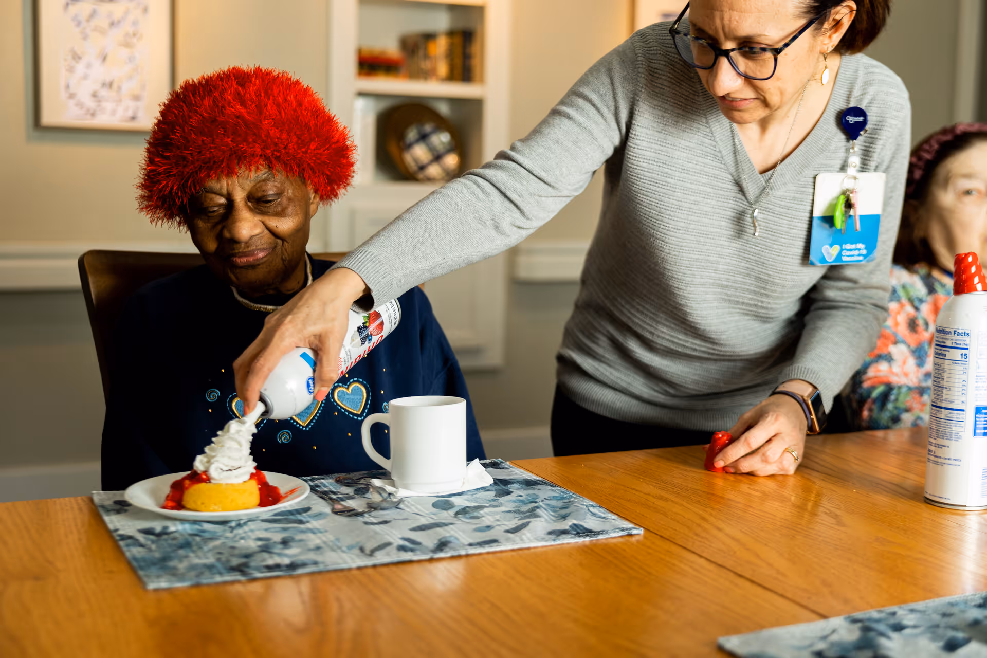 A caregiver sprays whipped cream onto a dessert for a seated elderly resident wearing a red hat at a dining table.