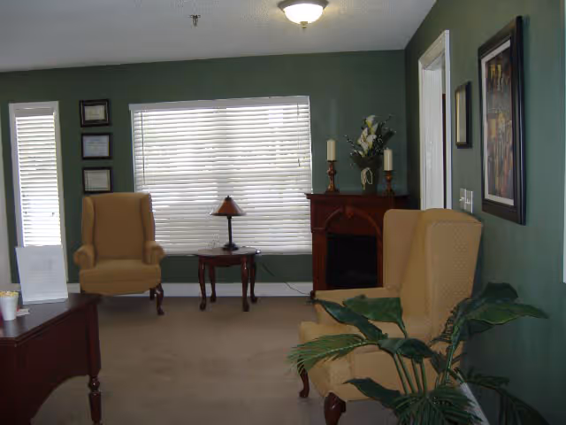 A cozy living room with green walls, two beige upholstered armchairs, a small wooden table with a lamp, a wooden cabinet with decorative candles and flowers, a large window with white blinds, framed pictures on the walls, and a potted plant in the foreground.