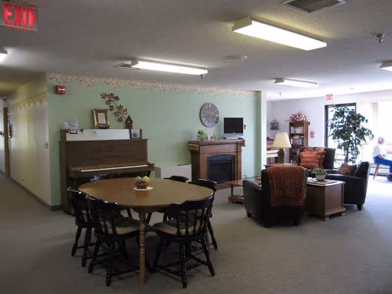 Common lounge area with round dining table, piano, fireplace, and seating in a senior residence.