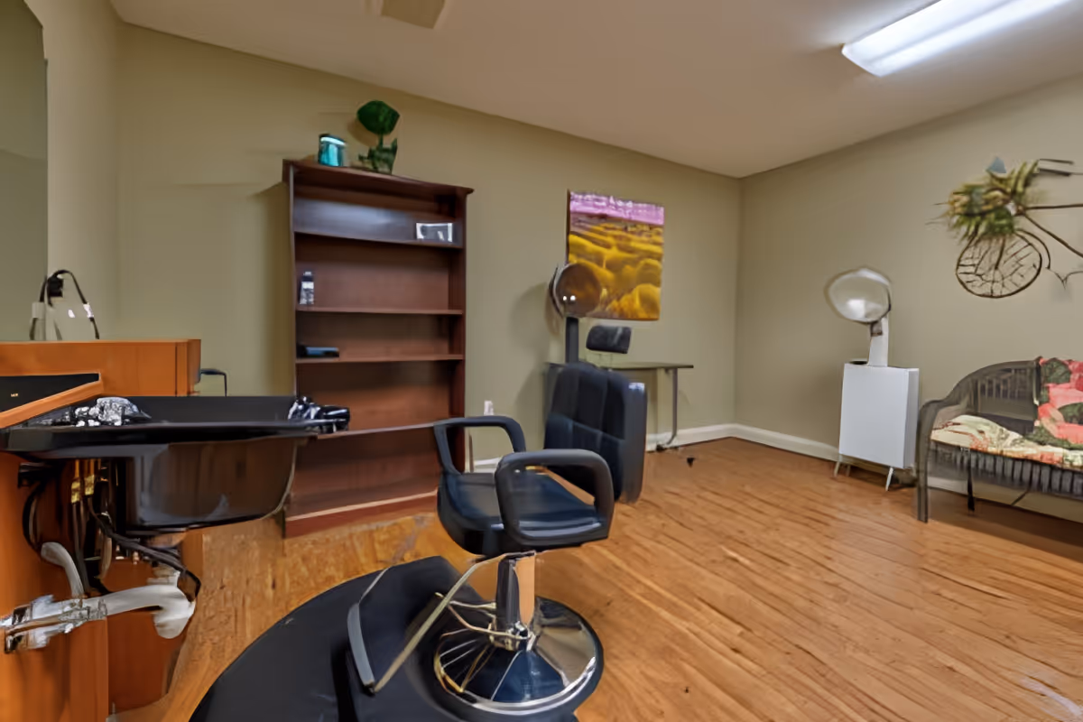 Interior of a hair salon area with a black salon chair in front of a hair washing station, a wooden bookshelf, a hair dryer chair, and a wicker bench with colorful cushions against beige walls and wooden flooring.