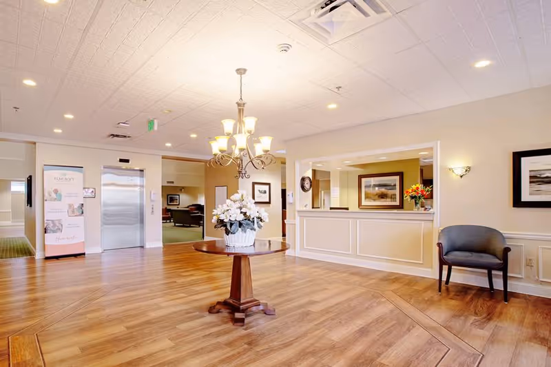 Bright assisted living lobby with a central round table holding a floral arrangement, a reception desk, elevator and seating.