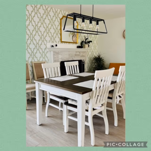 A dining room with a wooden table and six white chairs. The table has placemats and is positioned in front of a fireplace with a decorative mirror above it. A modern rectangular light fixture hangs above the table. The walls have a patterned wallpaper on one side and are plain white on the other. There are plants and additional chairs in the background.