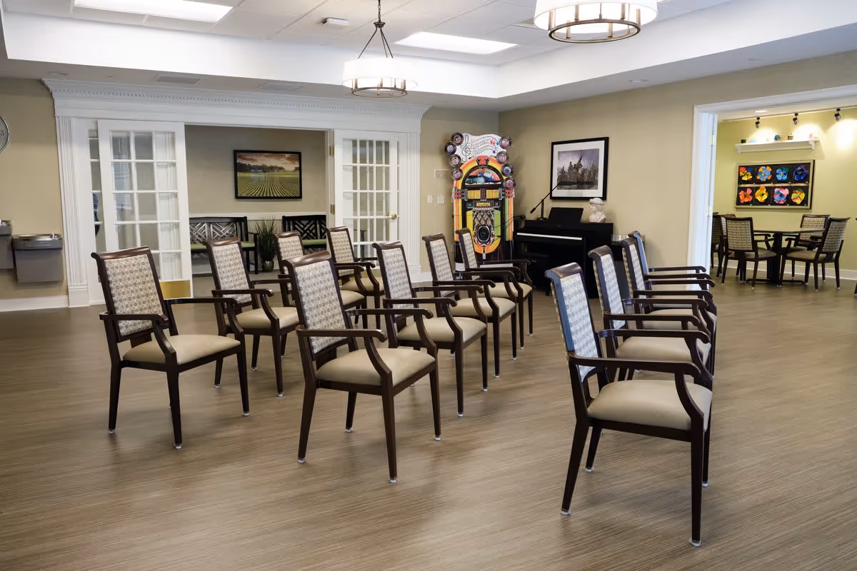 A room with two rows of wooden chairs with cushioned seats and backs arranged facing forward. In the background, there is a colorful jukebox, a black piano with a bust sculpture on top, and framed artwork on the walls. Through an open doorway, another room with tables and chairs and colorful artwork on the wall is visible. The floor is wood-patterned, and the ceiling has recessed lighting and two circular hanging light fixtures.