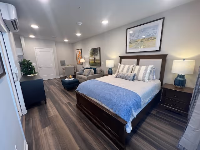 Furnished bedroom studio with a bed, two nightstands, a seating area, and wood-look floors.