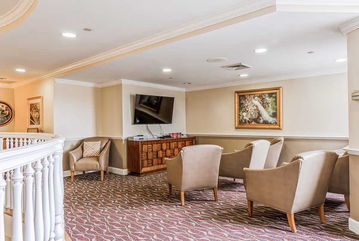 A cozy living room area with five beige armchairs arranged around a wooden cabinet with a flat-screen TV mounted above it. The room has patterned carpet flooring, beige walls with white trim, recessed ceiling lights, and framed artwork on the walls. A white railing is visible on the left side.