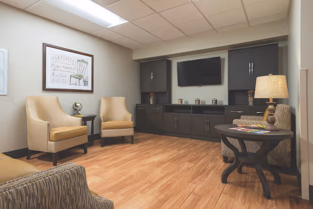 A cozy sitting area with armchairs, a round side table and lamp, built-in cabinetry with a wall-mounted TV, and wood flooring.