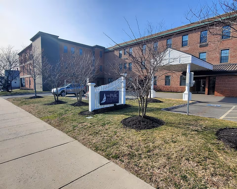 Exterior view of The Parc at Harbor View Senior Living facility showing a three-story brick building with multiple windows, a white picket fence sign with the facility name, leafless trees, a sidewalk, and a covered entrance with a parking area.
