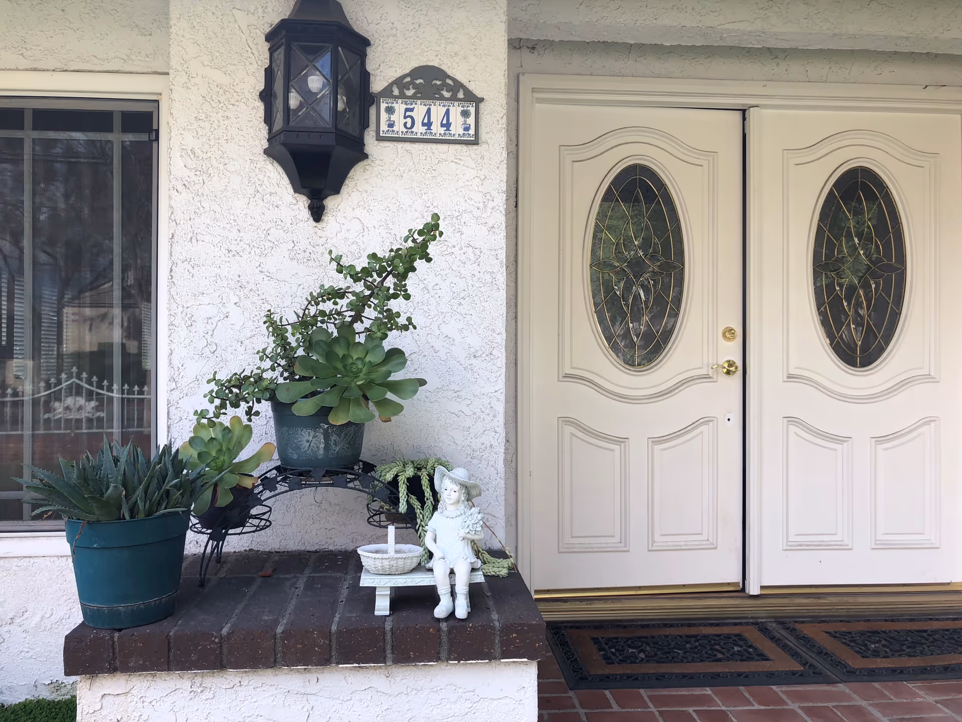 Front entrance of a building with double white doors featuring decorative oval glass panels. To the left of the doors is a small brick ledge with potted succulent plants and a small white statue of a girl sitting on a bench. Above the ledge is a black outdoor lantern light and a decorative house number plaque displaying the number 544.