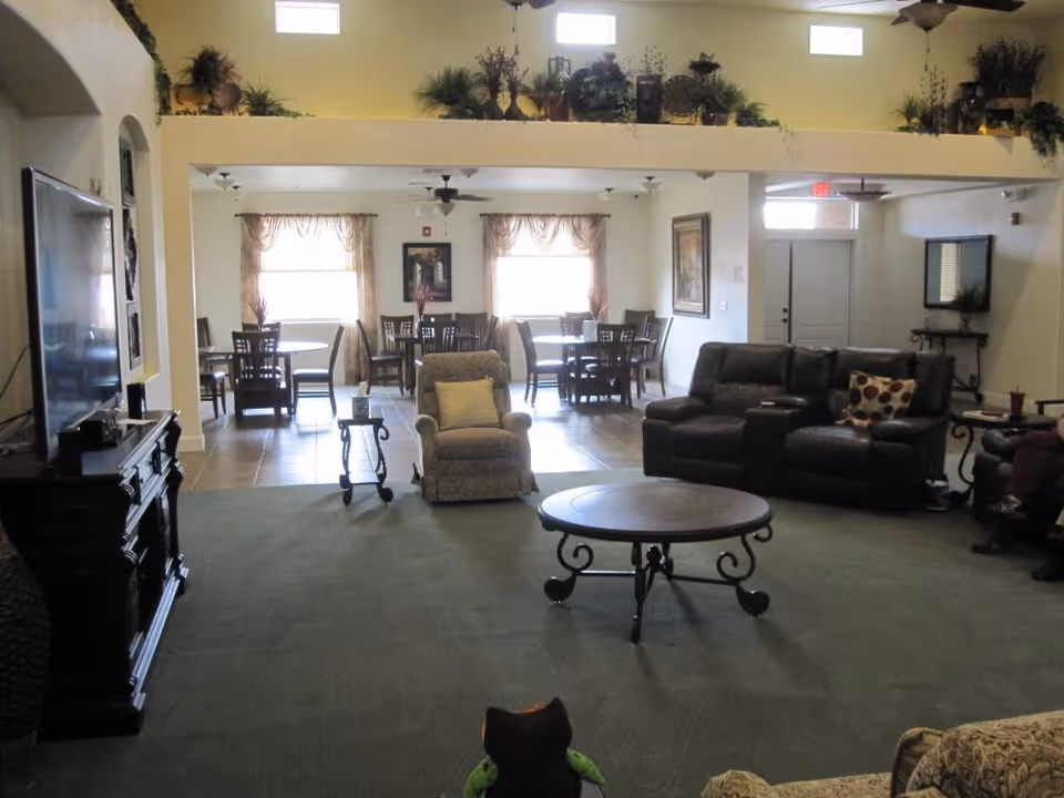 Spacious common living area with sofas, armchairs, a round coffee table and dining tables in the background under high windows.