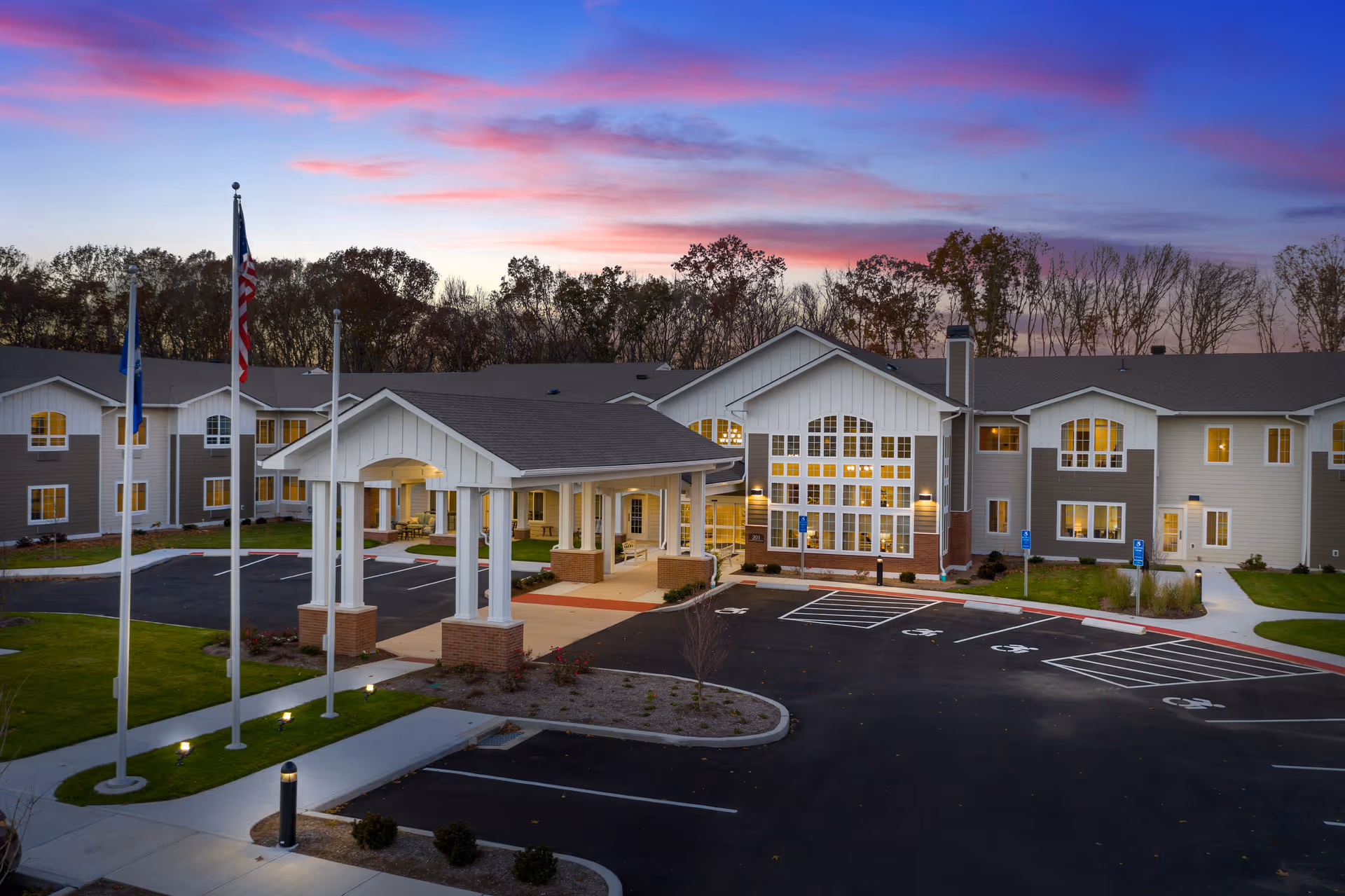 Exterior view of The Landing of North Haven senior living facility at dusk, showing a large two-story building with a covered entrance, multiple windows lit from inside, three flagpoles with flags, a parking lot with handicap spaces, and trees in the background under a colorful sky.