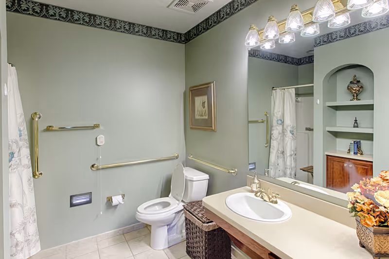 A clean and well-lit bathroom with light green walls and beige tiled floor. The bathroom features a white toilet with the lid open, a white sink with a beige countertop, and a large mirror above the sink with multiple light fixtures. There are gold-colored grab bars on the walls near the toilet and shower area. A shower curtain with a floral pattern is partially visible on the left side. A wicker basket is placed next to the toilet, and a framed picture hangs on the wall above the toilet. Built-in shelves with decorative items and a cabinet are visible in the reflection of the mirror.