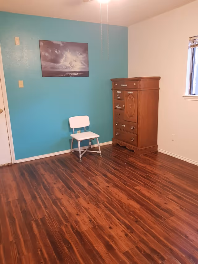 A room with wooden flooring, a teal accent wall, a white chair, a wooden dresser with labeled drawers, a window with blinds, and a ceiling fan with pull chains.
