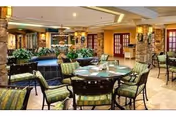 A spacious and well-lit common area with multiple round tables and cushioned chairs arranged around them. The room features stone pillars, indoor plants, and warm lighting, creating a cozy and inviting atmosphere.