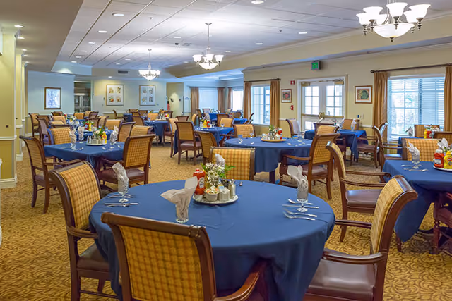 A spacious dining room with multiple round tables covered in blue tablecloths, each set with napkins, silverware, and condiments. The room has beige walls, carpeted floors, and several windows letting in natural light. Comfortable wooden chairs with cushioned seats surround the tables, and chandeliers hang from the ceiling providing warm lighting.