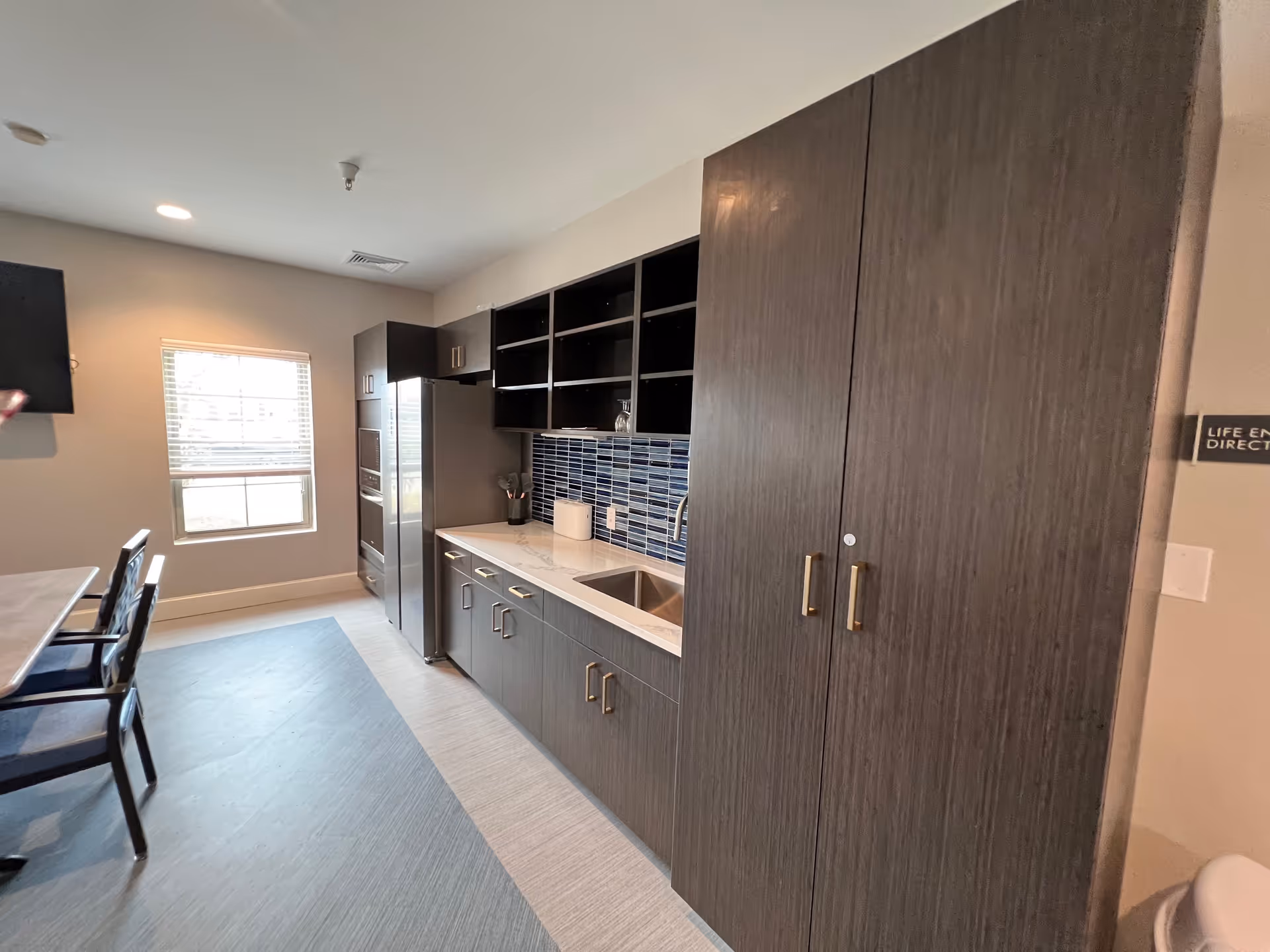 Modern kitchen area with dark wood cabinets, a stainless steel refrigerator, a sink with a blue tiled backsplash, and a window letting in natural light. There is a dining table with chairs on the left side of the image.