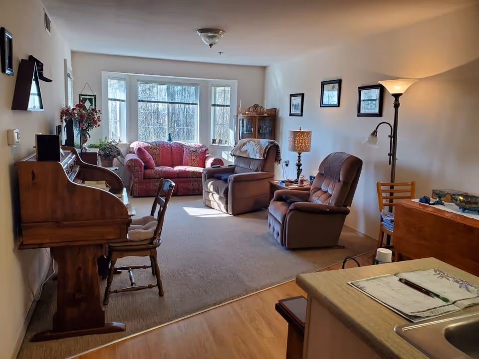 Sunlit living room with a red sofa and two recliners, a wooden desk and chairs, and a bay window.