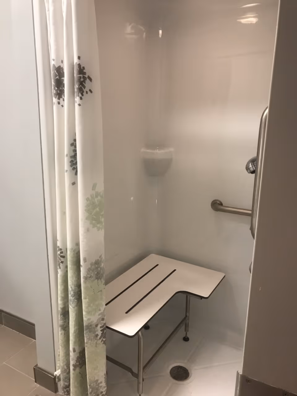 Accessible shower area with a white shower bench, grab bar, and a shower curtain with a floral pattern in shades of green and gray.