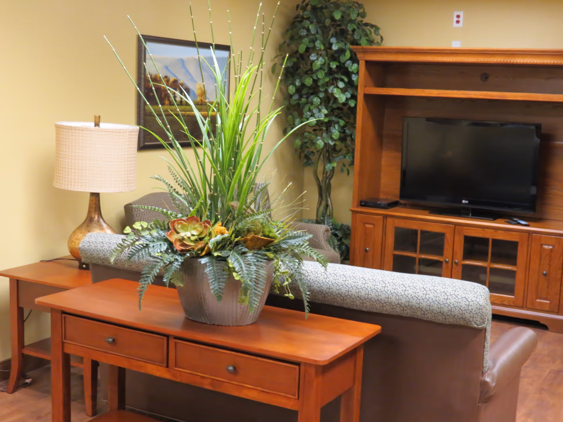 A cozy living room area with a wooden console table holding a large decorative plant. Behind the table is a cushioned armchair and a wooden entertainment center with a flat-screen TV. A table lamp and a framed landscape painting are visible on the left side, with a tall artificial plant in the corner.