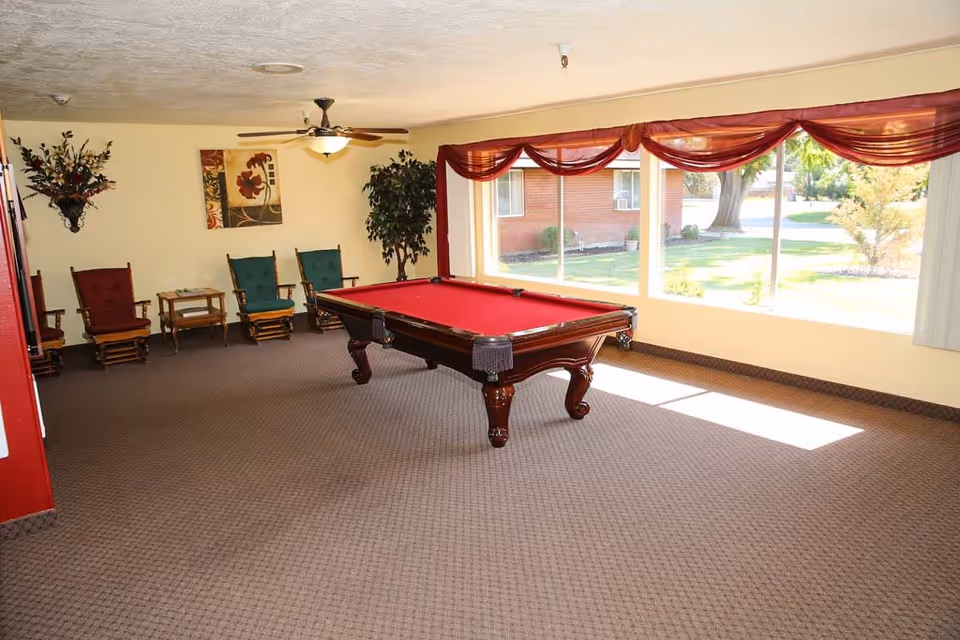 A bright room with a red pool table in the center, surrounded by several wooden rocking chairs with green and red cushions. The room has large windows with red curtains, allowing natural light to fill the space. There is a ceiling fan with a light fixture, a potted plant, and wall decorations including a floral painting and a wall-mounted flower arrangement.