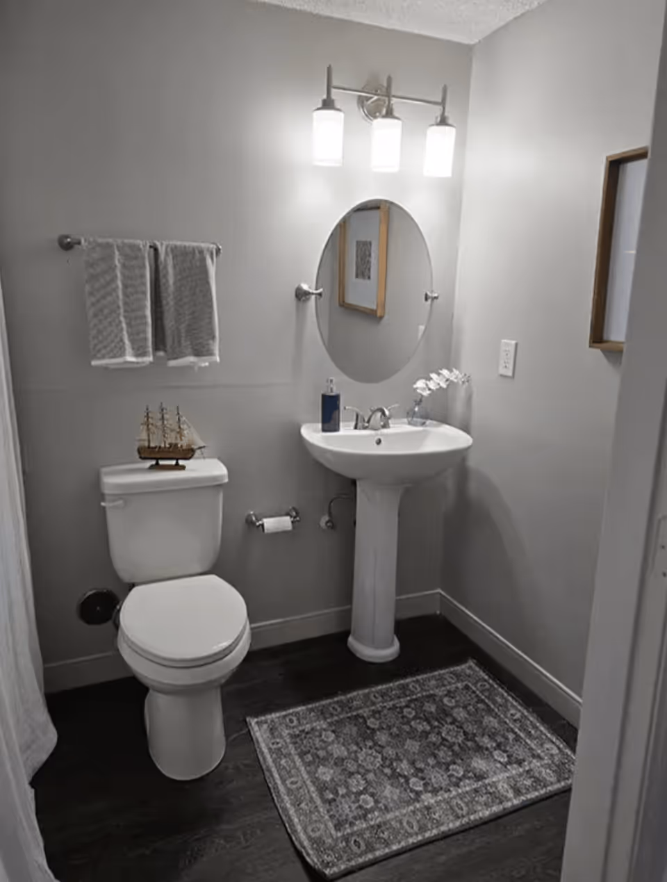 A small bathroom with a white toilet and pedestal sink. Above the sink is an oval mirror with three light fixtures. Two gray towels hang on a towel rack above the toilet, which has a decorative model ship on the tank. A patterned rug is on the dark floor, and a framed picture is partially visible on the wall.