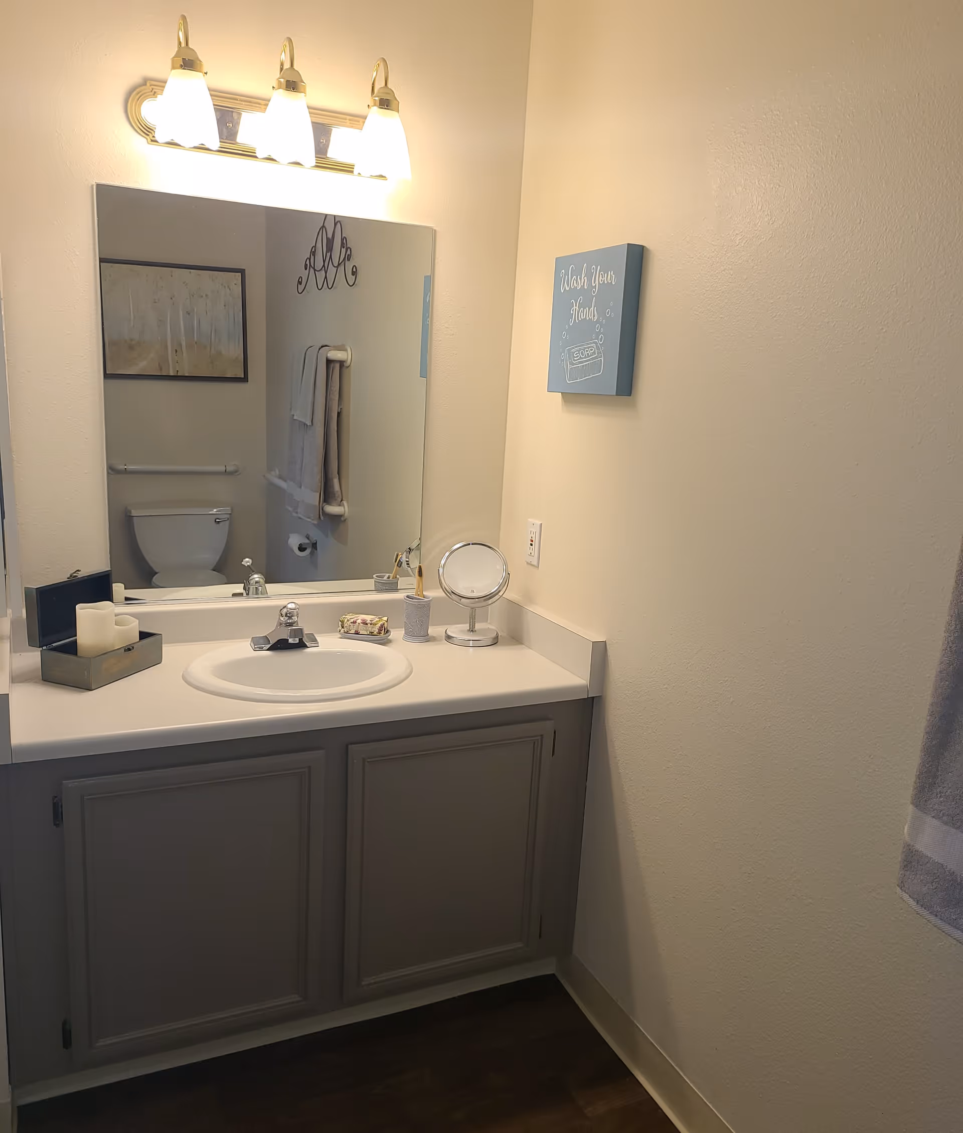 A clean bathroom vanity with a sink, a large mirror above it, and three light fixtures mounted on the wall. On the countertop, there are candles, a small mirror, a soap dish, and a toothbrush holder. In the mirror's reflection, a toilet, a towel rack with towels, and a framed picture are visible. A small wall decoration with the text 'Wash Your Hands' is mounted on the adjacent wall.