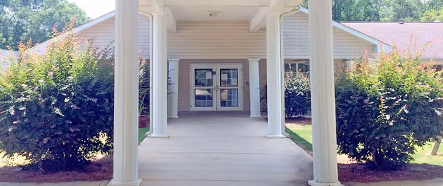 Covered entrance walkway with white columns leading to double glass doors and manicured shrubs on either side.