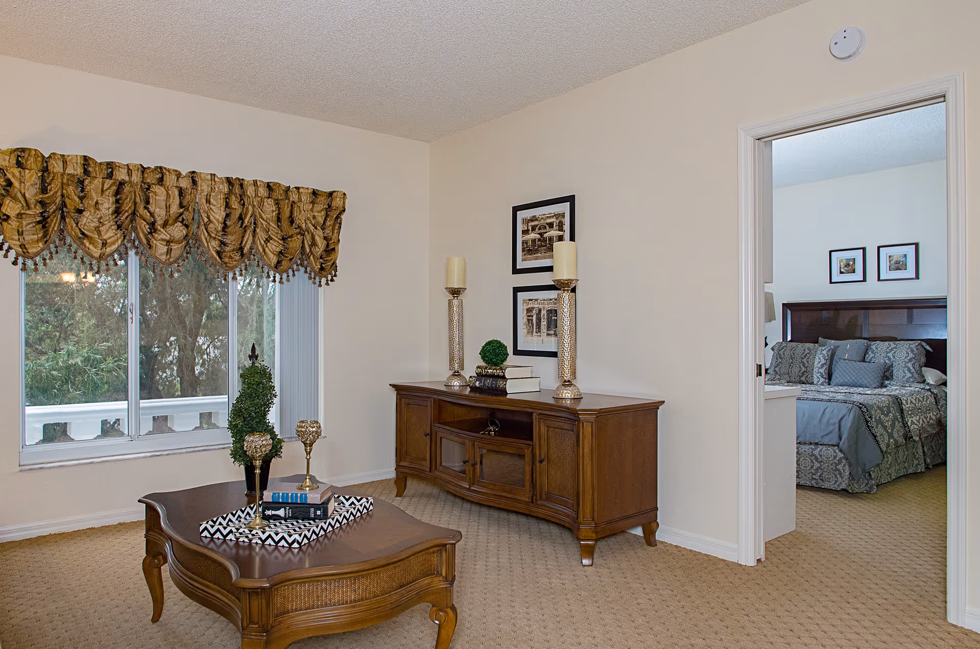 Bright furnished living room with a wooden coffee table and media console, window with a decorative valance, and a view into an adjacent bedroom.