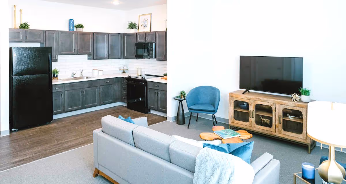 A modern open-concept living space featuring a kitchen with dark gray cabinets, black refrigerator, stove, and microwave. Adjacent to the kitchen is a living room area with a light gray sofa, a blue armchair, a wooden TV stand with a flat-screen TV, and a small round side table with a lamp. The floor transitions from wood in the kitchen to carpet in the living area.