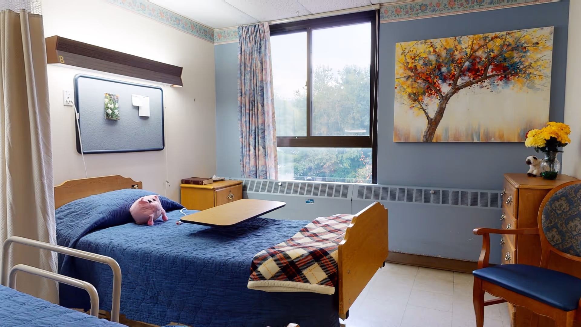 A neatly furnished senior care bedroom with a single bed covered in blue bedding, an overbed table, window, dresser and chair.
