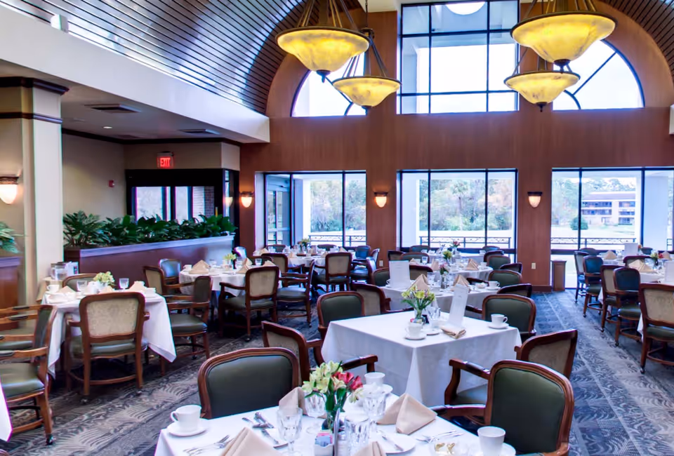 A bright and spacious dining room with multiple tables covered in white tablecloths, set with cups, glasses, napkins, and cutlery. The room features large windows allowing natural light to fill the space, elegant hanging light fixtures, and green cushioned chairs around each table. There are plants along a divider and a view of trees and buildings outside through the windows.
